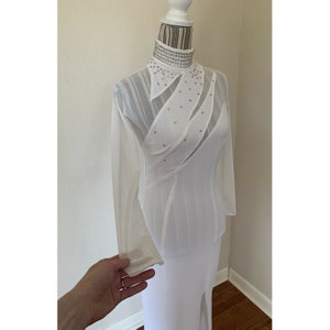 May include: A white, long-sleeved dress with a high collar and a side slit. The collar and bodice are adorned with sparkling embellishments. The dress is displayed on a mannequin.