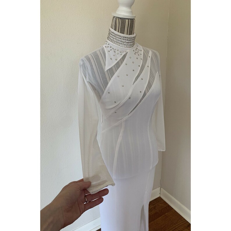 May include: A white, long-sleeved dress with a high collar and a side slit. The collar and bodice are adorned with sparkling embellishments. The dress is displayed on a mannequin.