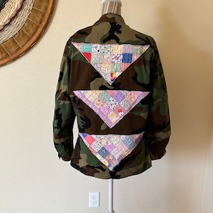 May include: A camouflage jacket with three colorful patchwork triangle accents on the back. The patches feature a variety of floral and patterned fabrics in shades of red, blue, purple, and yellow. The jacket is displayed on a white mannequin.