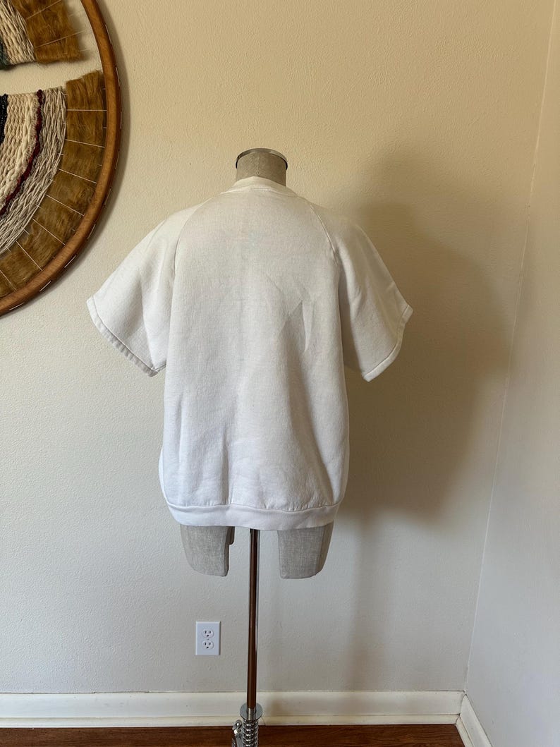 Op de afbeelding: Een witte sweatshirt met korte mouwen, getoond op een mannequin. De sweatshirt heeft een ronde hals en een relaxte pasvorm. De mouwen zijn kort en afgewerkt met een geribde boord. Het kledingstuk is gemaakt van zacht materiaal, waarschijnlijk katoen.