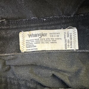 May include: Close-up of a pair of black Wrangler jeans. The tag reads "Wrangler" and includes washing instructions and size information. The jeans are made of 100% cotton.
