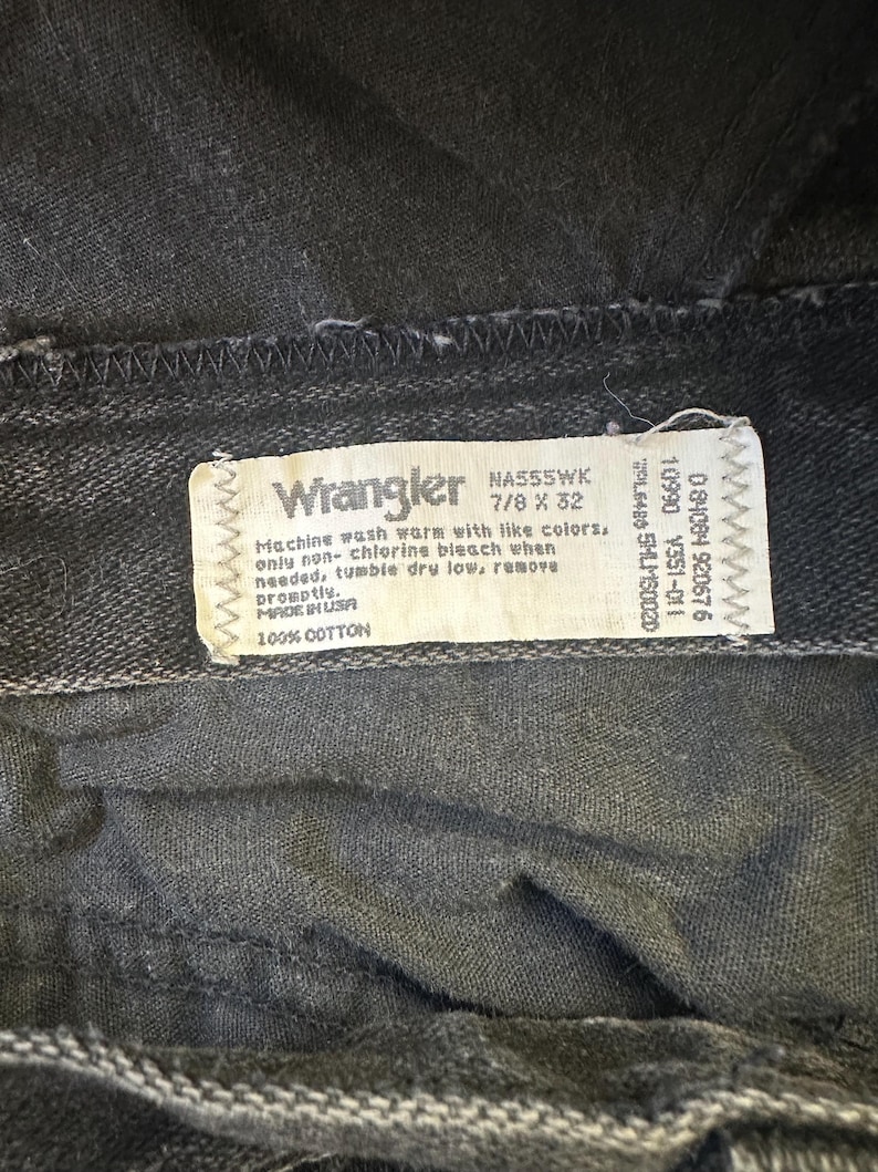 May include: Close-up of a pair of black Wrangler jeans. The tag reads "Wrangler" and includes washing instructions and size information. The jeans are made of 100% cotton.