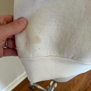 Op de afbeelding: Cr&egrave;mekleurige sweatshirt met geribde zoom en manchetten. De stof vertoont een vlek en enkele kleine donkere vlekken. Het kledingstuk wordt vastgehouden door een hand met een zilveren ring.