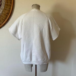 Op de afbeelding: Een witte sweatshirt met korte mouwen, gepresenteerd op een mannequin. Het kledingstuk heeft een ronde hals en een geribbelde zoom. De mouwen zijn kort en wijd. Het materiaal lijkt zacht te zijn, waarschijnlijk katoen.