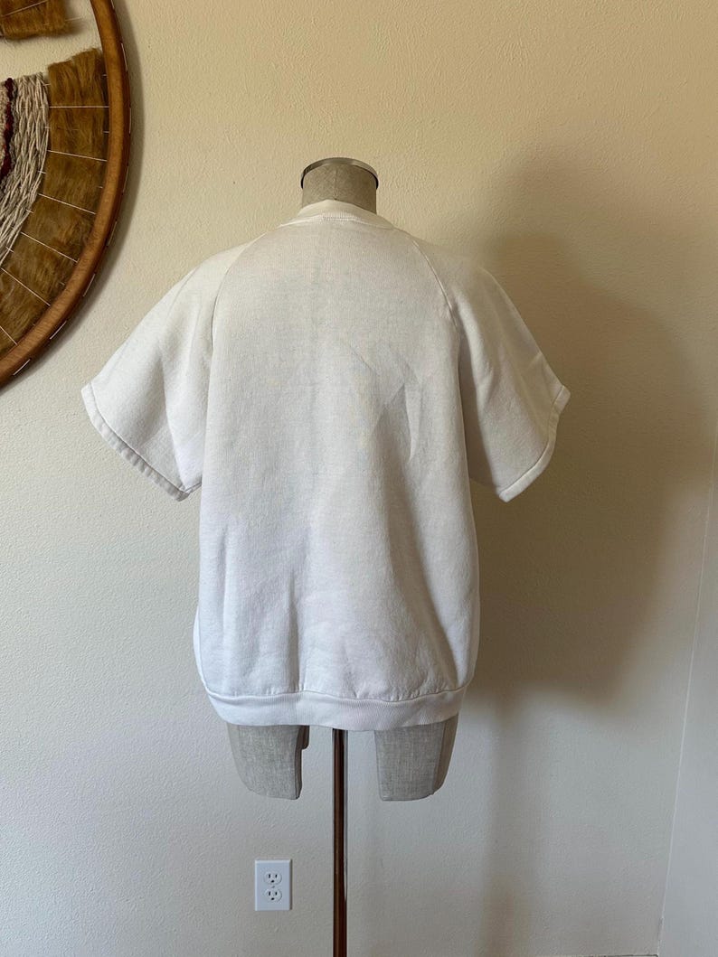Op de afbeelding: Een witte sweatshirt met korte mouwen, gepresenteerd op een mannequin. Het kledingstuk heeft een ronde hals en een geribbelde zoom. De mouwen zijn kort en wijd. Het materiaal lijkt zacht te zijn, waarschijnlijk katoen.