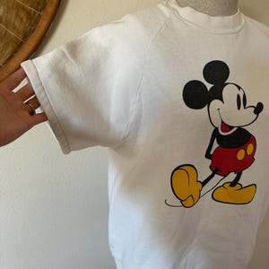 Op de afbeelding: Witte sweatshirt met een Mickey Mouse-afbeelding. Het stripfiguur is afgebeeld in zijn iconische outfit: rode shorts, gele schoenen en zwarte handschoenen. De sweatshirt heeft een ronde hals en raglanmouwen.