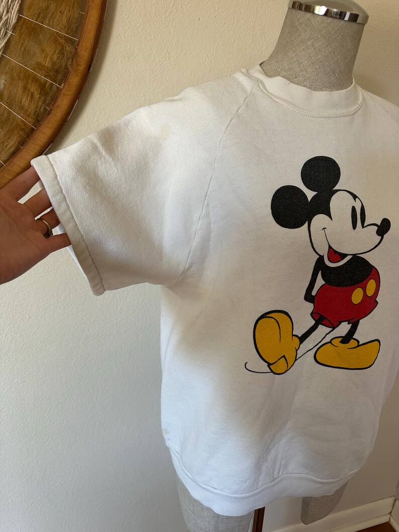 Op de afbeelding: Witte sweatshirt met een Mickey Mouse-afbeelding. Het stripfiguur is afgebeeld in zijn iconische outfit: rode shorts, gele schoenen en zwarte handschoenen. De sweatshirt heeft een ronde hals en raglanmouwen.