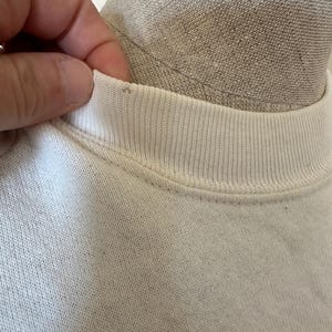 Op de afbeelding: Close-up van een witte sweatshirt met een geribbelde ronde hals. De stof lijkt licht versleten. Een hand raakt de kraag van het shirt aan.
