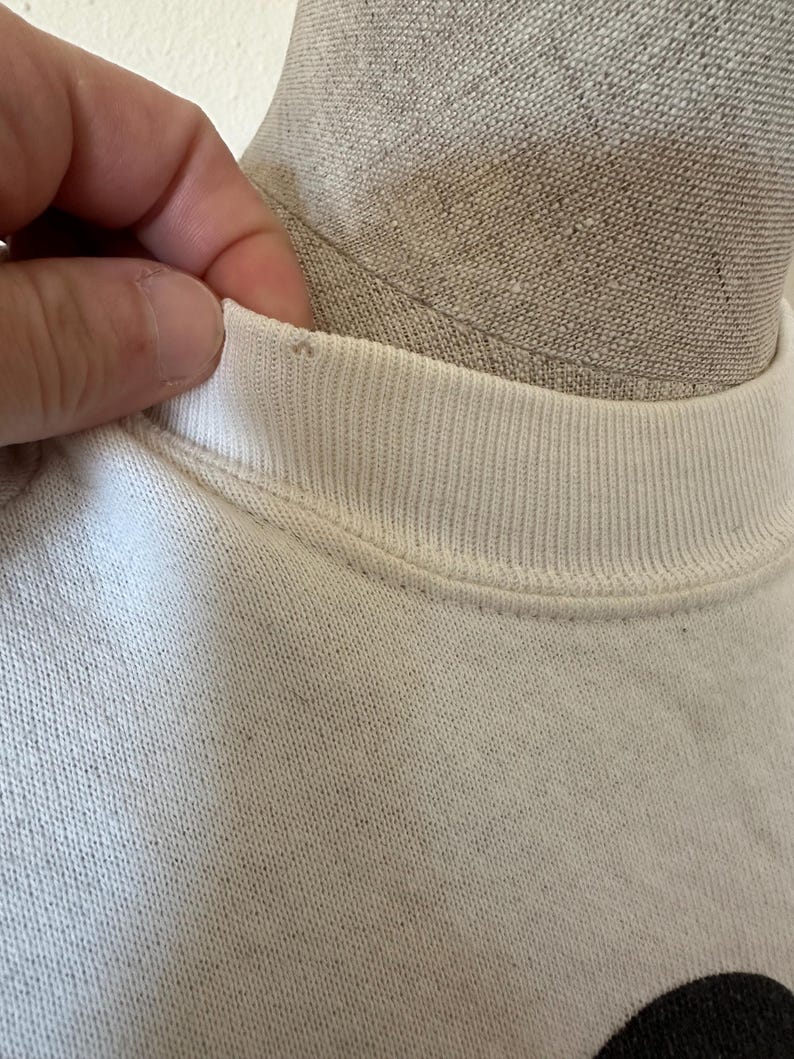 Op de afbeelding: Close-up van een witte sweatshirt met een geribbelde ronde hals. De stof lijkt licht versleten. Een hand raakt de kraag van het shirt aan.