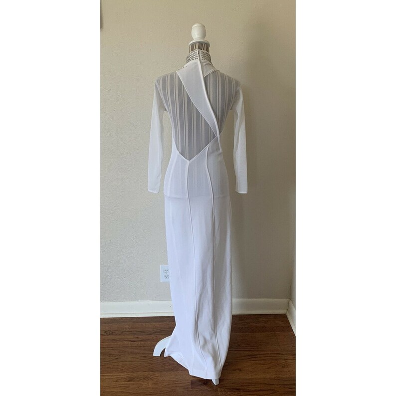 May include: A white, floor-length gown with long sleeves and a distinctive open back design. The dress has a high neckline and a sheer, vertically striped panel across the upper back. The dress is displayed on a mannequin.