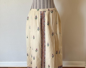 May include: Cream-coloured skirt with a floral pattern. The skirt has a central vertical panel with a detailed border design and is decorated with small floral motifs. The skirt is displayed on a mannequin.