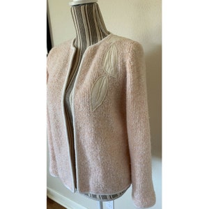 May include: A light pink open-front jacket with a textured knit fabric. The jacket features a cream-colored trim and two leaf-shaped appliqués on the upper left side. The appliqués are embellished with small, white beads.