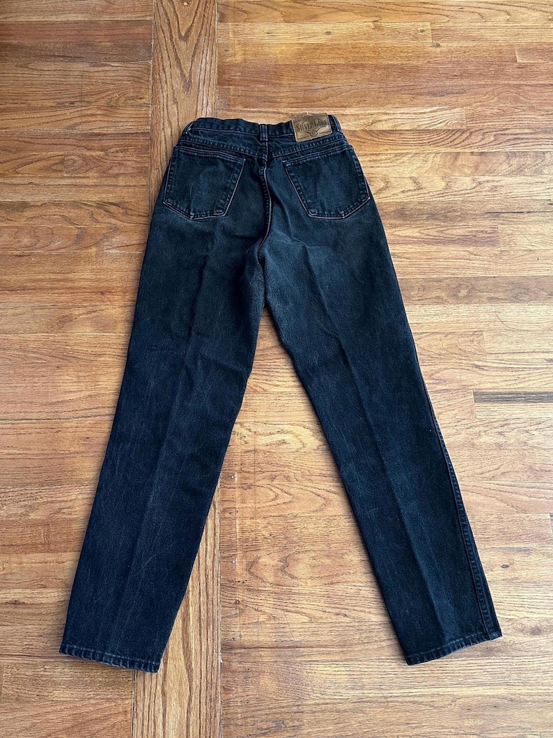 May include: Dark wash denim jeans with a classic five-pocket design. The jeans have a straight leg cut and a faded appearance, with a leather patch on the back waistband. The jeans are laid flat on a wooden surface.