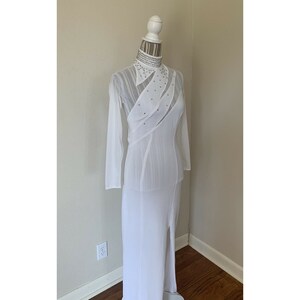 May include: A white, floor-length gown with long sleeves and a high collar. The dress features a sheer overlay with rhinestone accents and a thigh-high slit. The dress is displayed on a mannequin.
