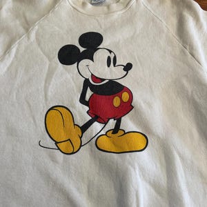 Op de afbeelding: Cr&egrave;mekleurige sweatshirt met een Mickey Mouse-afbeelding. Mickey wordt getoond in zijn iconische pose, met een rode broek, gele schoenen en zwarte handschoenen. De sweatshirt heeft een ronde hals en raglanmouwen.