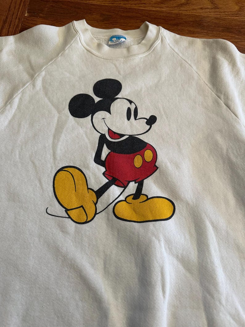 Op de afbeelding: Cr&egrave;mekleurige sweatshirt met een Mickey Mouse-afbeelding. Mickey wordt getoond in zijn iconische pose, met een rode broek, gele schoenen en zwarte handschoenen. De sweatshirt heeft een ronde hals en raglanmouwen.