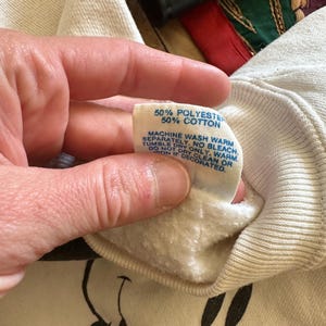Op de afbeelding: Cr&egrave;mekleurige sweatshirt met een Mickey Mouse-afbeelding. Het label luidt "50% Polyester 50% Katoen" en bevat wasvoorschriften. De sweatshirt wordt door een persoon vastgehouden.