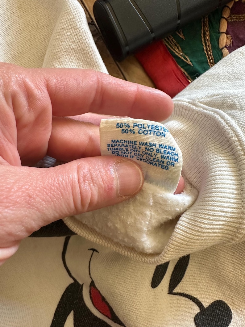 Op de afbeelding: Cr&egrave;mekleurige sweatshirt met een Mickey Mouse-afbeelding. Het label luidt "50% Polyester 50% Katoen" en bevat wasvoorschriften. De sweatshirt wordt door een persoon vastgehouden.