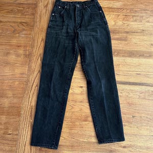 May include: Black denim jeans with a classic five-pocket design. The jeans have a high waist and a straight leg cut. The dark wash denim shows subtle fading and wear, giving them a vintage look. The jeans are laid flat on a wooden surface.