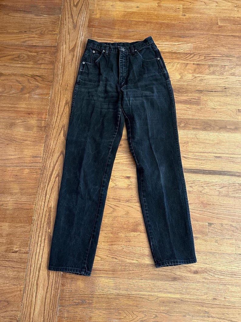 May include: Black denim jeans with a classic five-pocket design. The jeans have a high waist and a straight leg cut. The dark wash denim shows subtle fading and wear, giving them a vintage look. The jeans are laid flat on a wooden surface.
