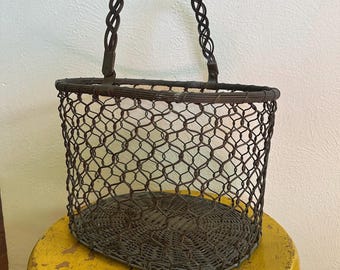 May include: A vintage wire basket with a woven handle. The basket has a rounded shape and a dark brown color. It is sitting on a yellow stool.