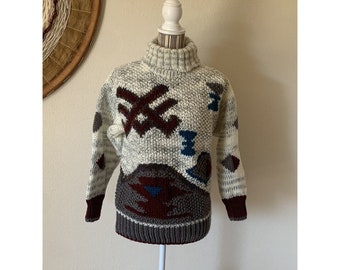 May include: A vintage, hand-knitted sweater with a turtleneck collar. The sweater features a cream-colored base with gray flecks and geometric patterns in burgundy, blue, and gray. The cuffs and hem are a darker gray. The sweater is displayed on a white mannequin.