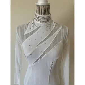 May include: White long-sleeved dress with a high, embellished collar. The dress features sheer panels and diagonal accents adorned with small, silver-coloured studs. The dress is displayed on a mannequin.