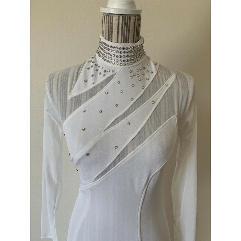 May include: White long-sleeved dress with a high, embellished collar. The dress features sheer panels and diagonal accents adorned with small, silver-coloured studs. The dress is displayed on a mannequin.