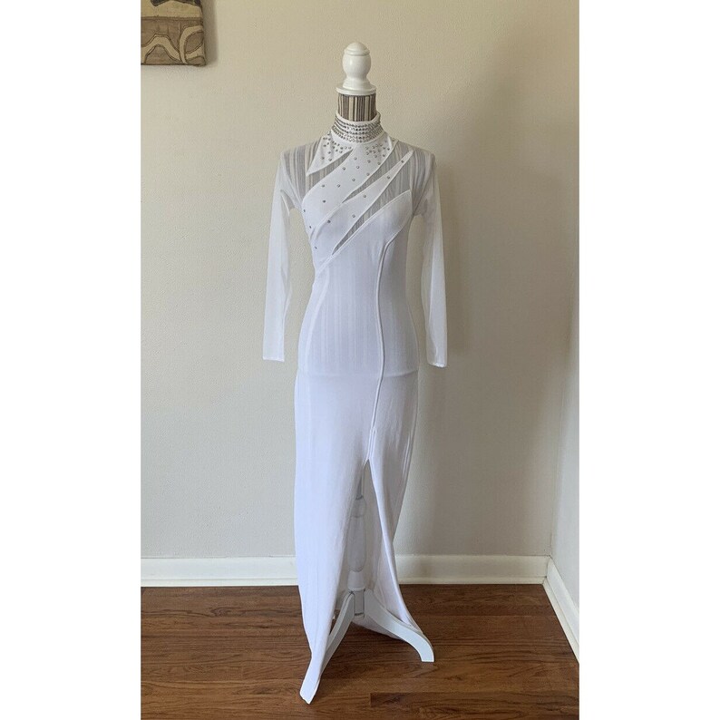 May include: A white, floor-length dress with long sleeves and a high neckline. The dress features sheer panels and is embellished with rhinestones. The dress has a slit up the front and is displayed on a mannequin.