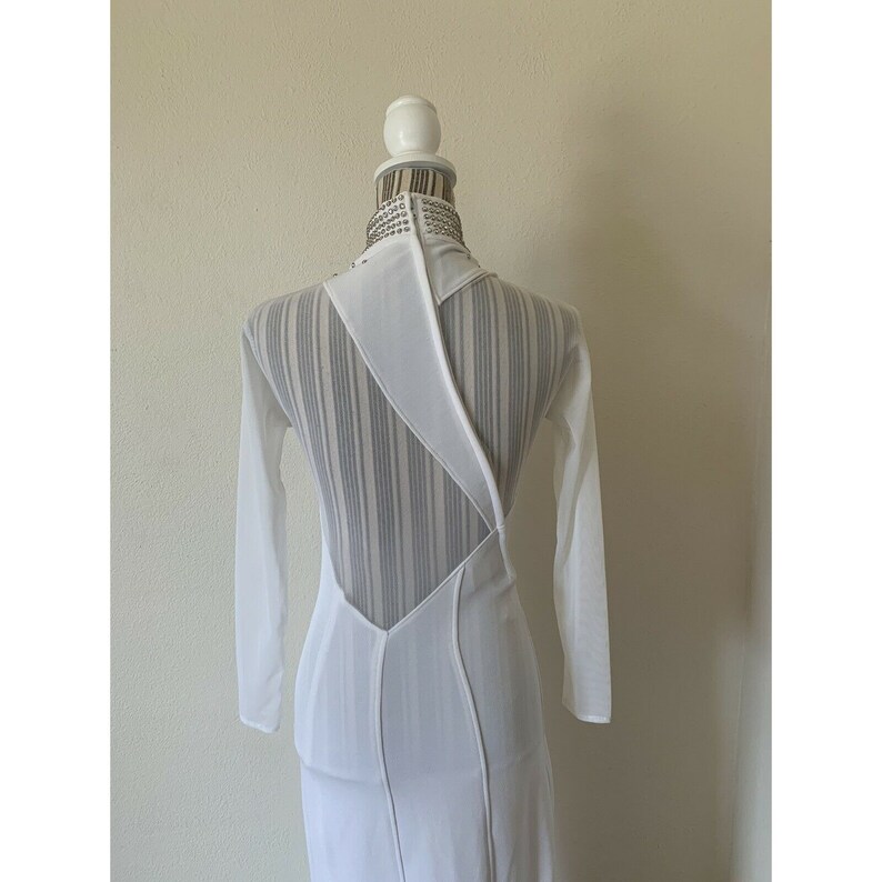 May include: White long-sleeved dress with a distinctive open back design. The dress has a high neckline with silver embellishments and a sheer back panel with vertical stripes. The dress has a fitted silhouette and a floor-length hemline.