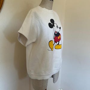 Op de afbeelding: Wit sweatshirt met korte mouwen met een Mickey Mouse-afbeelding. Het stripfiguur is afgebeeld in zijn klassieke outfit: zwarte oren, rode shorts en gele schoenen. Het sweatshirt wordt op een mannequin gepresenteerd.