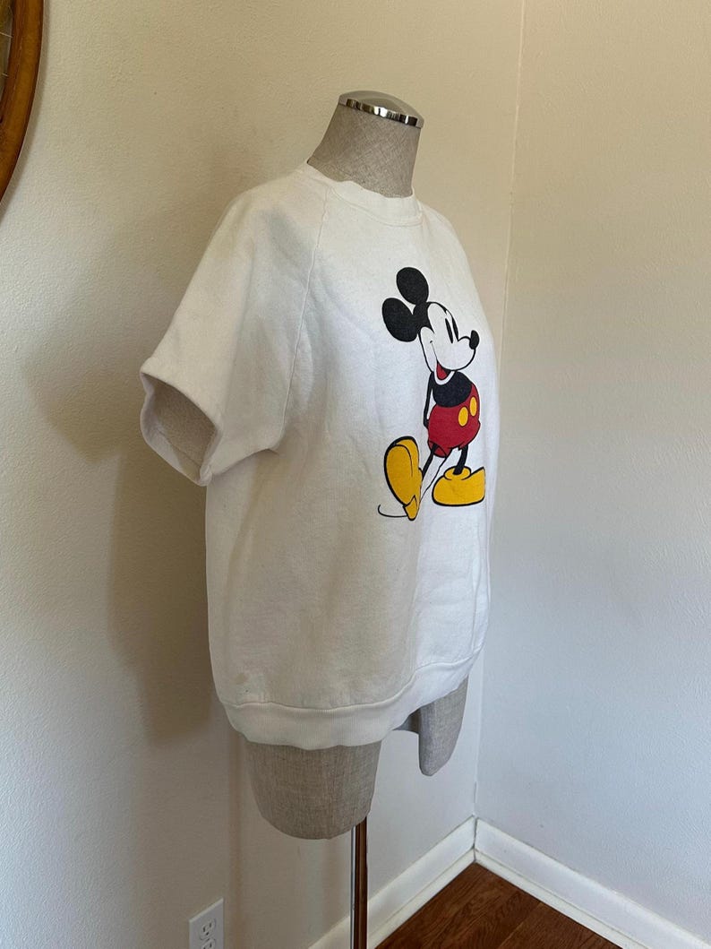 Op de afbeelding: Wit sweatshirt met korte mouwen met een Mickey Mouse-afbeelding. Het stripfiguur is afgebeeld in zijn klassieke outfit: zwarte oren, rode shorts en gele schoenen. Het sweatshirt wordt op een mannequin gepresenteerd.