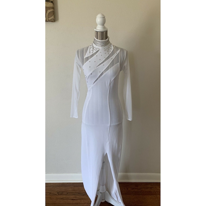May include: A white, long-sleeved dress with a high neckline and a front slit. The dress features sheer panels and is embellished with small, sparkling accents. The dress is displayed on a white mannequin.