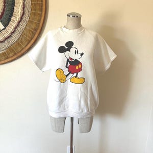 Op de afbeelding: Wit sweatshirt met korte mouwen met een Mickey Mouse-afbeelding. Het stripfiguur is in zwart, rood en geel. Het sweatshirt wordt op een mannequin getoond.