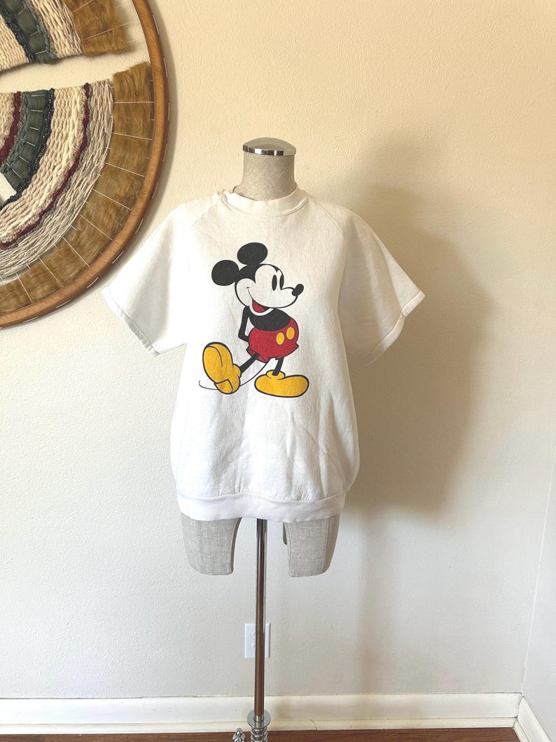 Op de afbeelding: Wit sweatshirt met korte mouwen met een Mickey Mouse-afbeelding. Het stripfiguur is in zwart, rood en geel. Het sweatshirt wordt op een mannequin getoond.