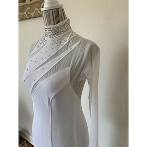 May include: White long-sleeved dress with a high, embellished collar. The dress features sheer mesh panels and diagonal fabric accents adorned with small, silver-coloured studs. The dress is displayed on a mannequin.