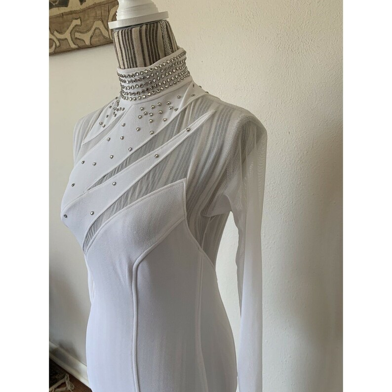 May include: White long-sleeved dress with a high, embellished collar. The dress features sheer mesh panels and diagonal fabric accents adorned with small, silver-coloured studs. The dress is displayed on a mannequin.