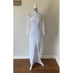 May include: A white, floor-length dress with long sleeves and a high neckline. The dress features sheer panels and a high slit on the left side. The bodice is embellished with small, sparkling accents. The dress is displayed on a white mannequin.