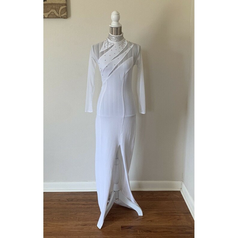 May include: A white, floor-length dress with long sleeves and a high neckline. The dress features sheer panels and a high slit on the left side. The bodice is embellished with small, sparkling accents. The dress is displayed on a white mannequin.