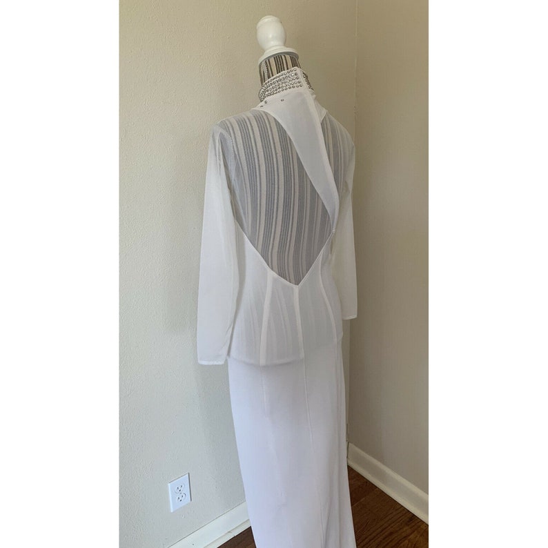 May include: A white formal dress with long sleeves and a striking open back design. The dress is displayed on a mannequin, highlighting its elegant silhouette and flowing fabric. The back features a sheer panel with vertical stripes.