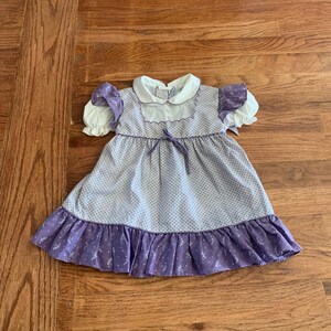 May include: A vintage baby dress in lavender and white. The dress features a white collar, short puffed sleeves, and a lavender bow at the waist. The skirt has a ruffled hem with a floral pattern. The dress is made of a soft, lightweight fabric.