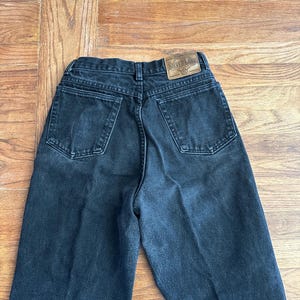 May include: Black denim jeans with a high waist and a classic five-pocket design. The jeans have a faded appearance and a leather patch with the text "Silver Lake" on the back waistband. The jeans are laid flat on a wooden surface.
