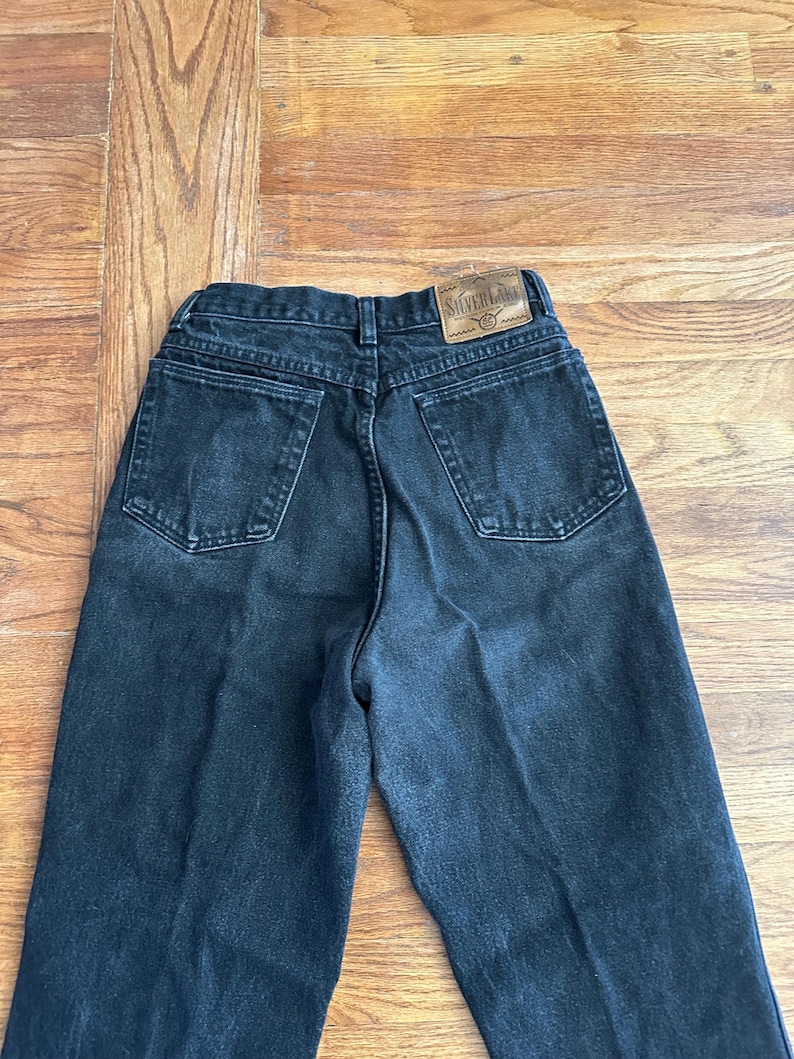 May include: Black denim jeans with a high waist and a classic five-pocket design. The jeans have a faded appearance and a leather patch with the text "Silver Lake" on the back waistband. The jeans are laid flat on a wooden surface.