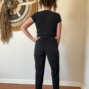 May include: A person wearing a black t-shirt and black denim jeans. The jeans have a classic, straight-leg fit. The person is standing in front of a wall with a woven wall hanging.