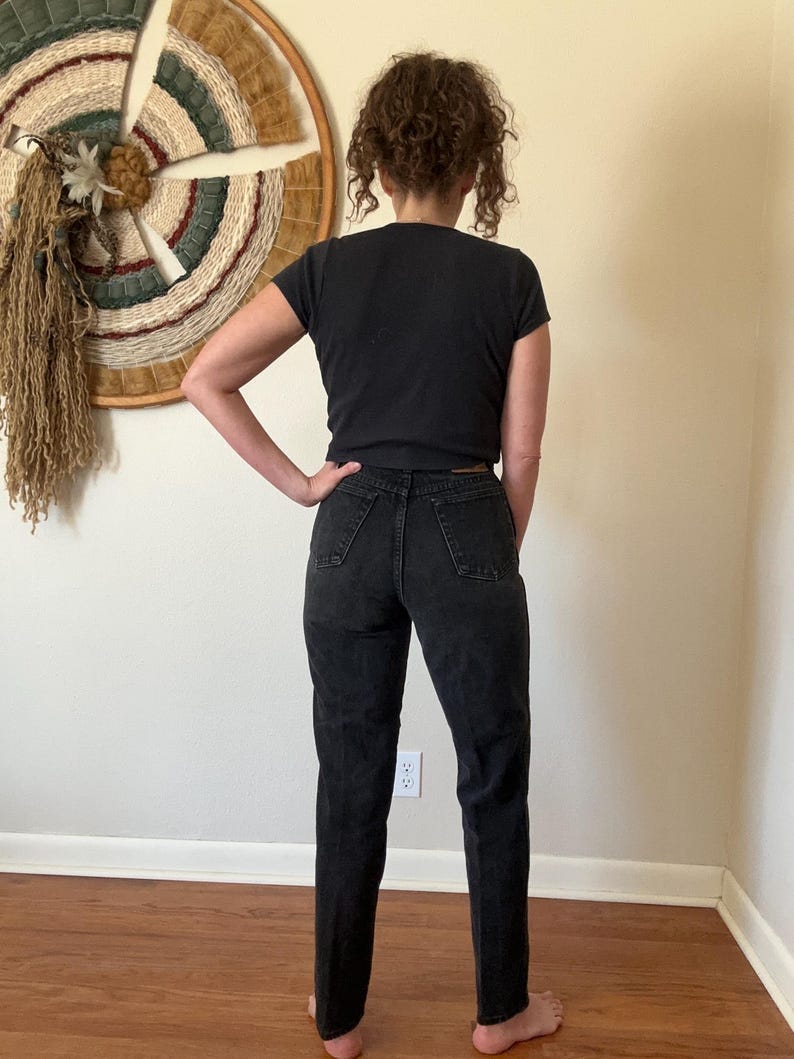 May include: A person wearing a black t-shirt and black denim jeans. The jeans have a classic, straight-leg fit. The person is standing in front of a wall with a woven wall hanging.