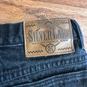 May include: Close-up of black denim jeans with a brown leather patch. The patch reads "Wrangler SILVERLAKE WESTERN FASHION JEANS" in a stylized font. The jeans have a classic five-pocket design.