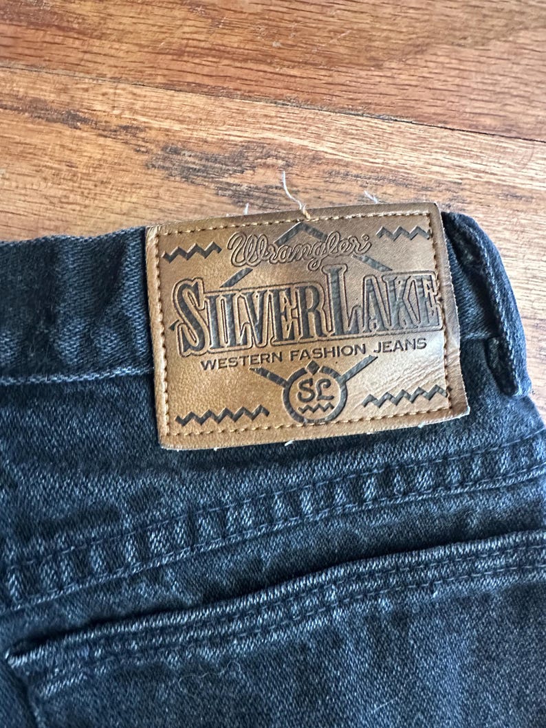 May include: Close-up of black denim jeans with a brown leather patch. The patch reads "Wrangler SILVERLAKE WESTERN FASHION JEANS" in a stylized font. The jeans have a classic five-pocket design.