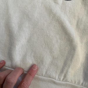 Op de afbeelding: Cr&egrave;mekleurige sweatshirt met een geel en zwart cartoonkarakterontwerp aan de bovenkant. De onderste zoom is geribbeld en de stof lijkt een zachte katoenmix te zijn. De afbeelding toont een close-up van de stof en het ontwerp.