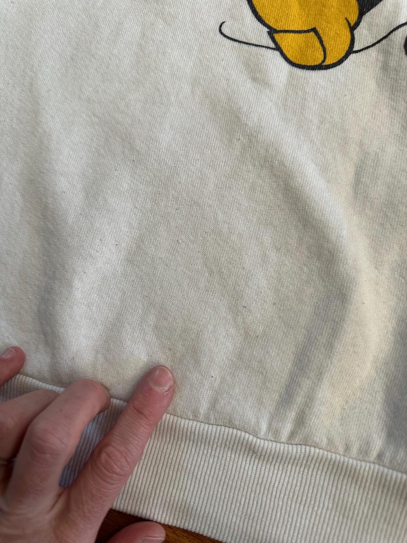 Op de afbeelding: Cr&egrave;mekleurige sweatshirt met een geel en zwart cartoonkarakterontwerp aan de bovenkant. De onderste zoom is geribbeld en de stof lijkt een zachte katoenmix te zijn. De afbeelding toont een close-up van de stof en het ontwerp.