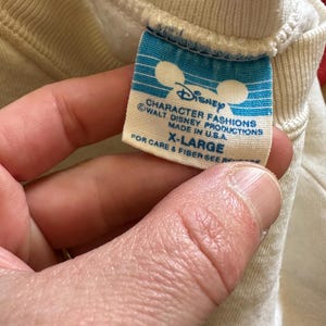 Op de afbeelding: Close-up van een blauw en wit Disney-label op een cr&egrave;mekleurig kledingstuk. Het label leest "CHARACTER FASHIONS" en "MADE IN U.S.A." met het Disney-logo en de tekst "X-LARGE". Het kledingstuk is gemaakt van een zachte, geribbelde stof.